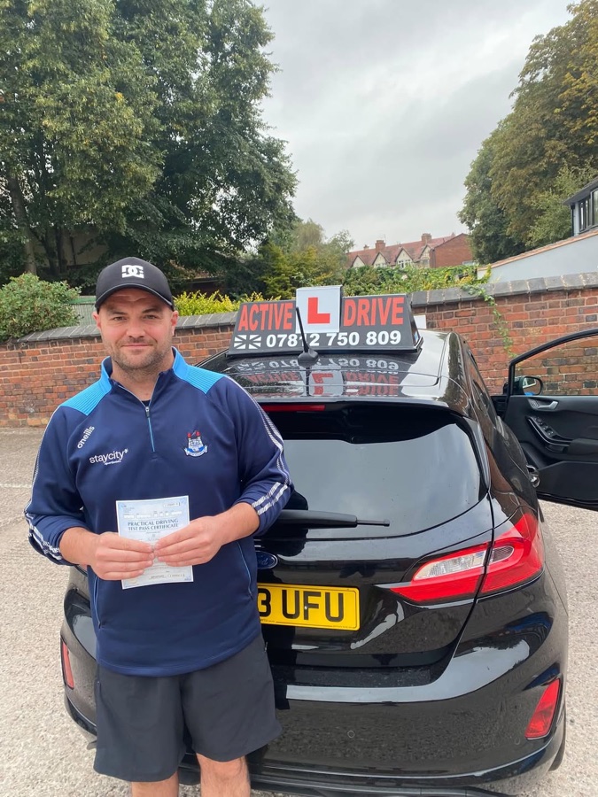 A real Active Drive student in a cap and navy zip-top holding his pass certificate beside the Active Drive car, with trees and terraced houses behind him