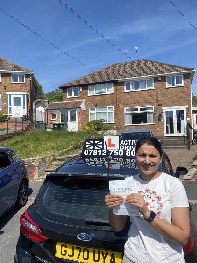 A real Active Drive student in a white t-shirt holding her pass certificate on a sunny street of semi-detached houses, beside the Active Drive car