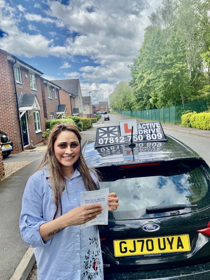 A real Active Drive student in a blue floral shirt holding her pass certificate on a residential street, beside the Active Drive car